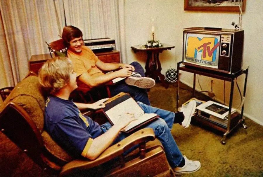 Two people sit in a living room watching MTV on a vintage box TV. One is holding a book and smiling, while the other sits nearby. The decor includes carpet, curtains, and retro furniture, evoking a 1980s vibe.