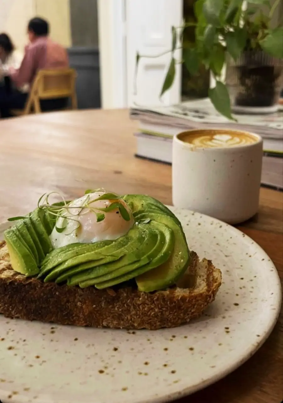 A plate with avocado toast topped with a poached egg and microgreens sits on a wooden table next to a cup of latte. In the background, people are seated and a potted plant is on the table.