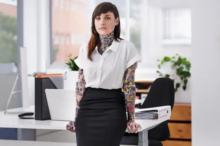 A woman with visible tattoos on her arms and neck, wearing a white blouse and black skirt, stands confidently by a desk in a modern office with bright natural light and plants in the background.