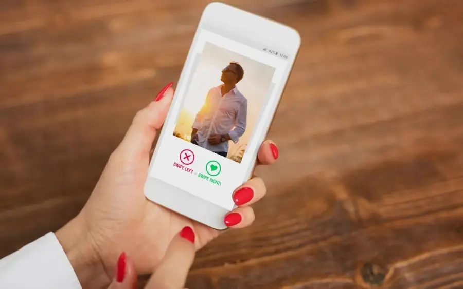 A person holding a smartphone displays a dating app profile with a man's photo, options to swipe left or right, and red-painted fingernails visible against a wooden table background.