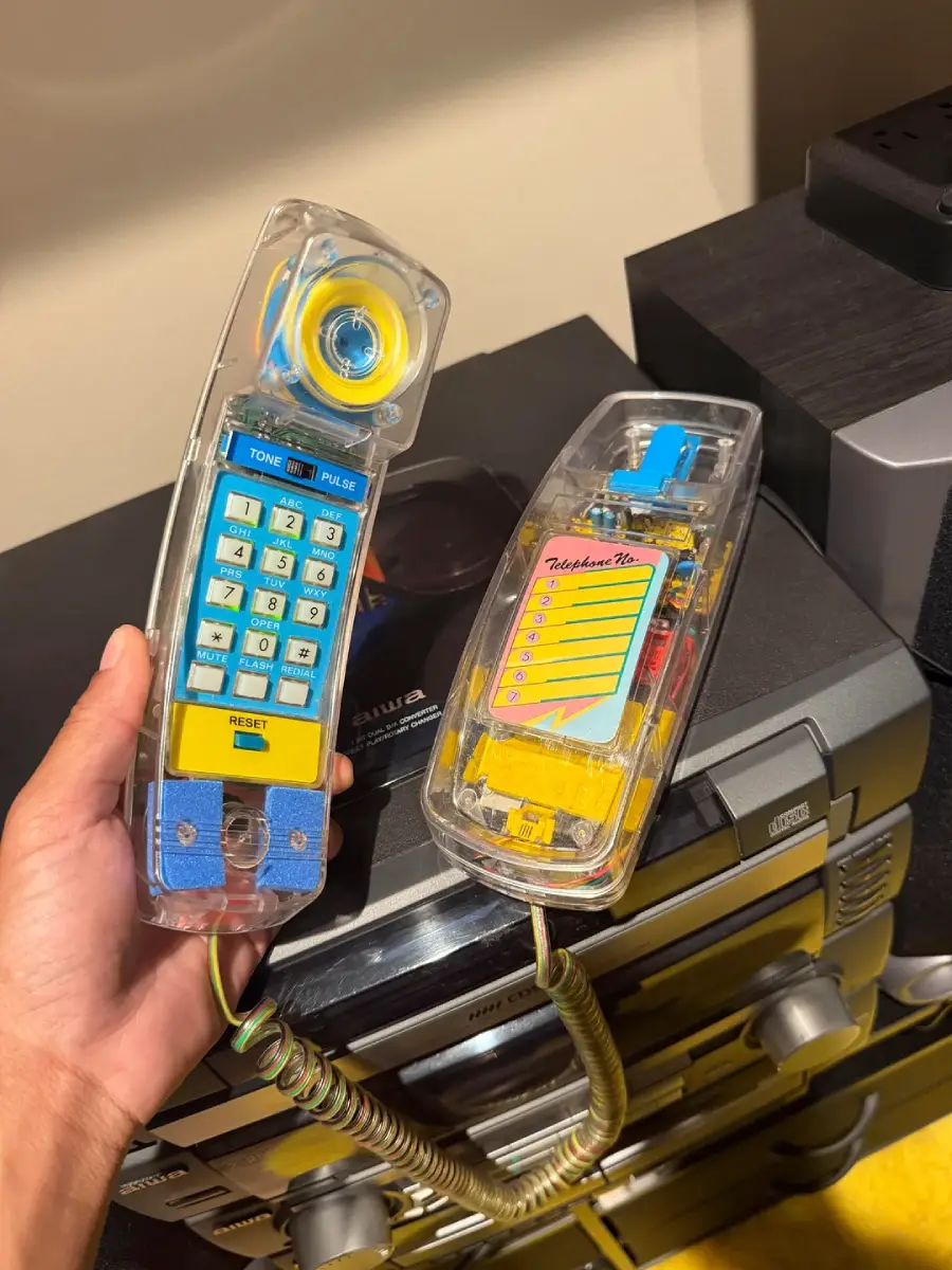 A hand holds a transparent landline phone with visible internal parts, blue buttons, and a coiled cord; the phone rests on a stereo system with speakers in the background.