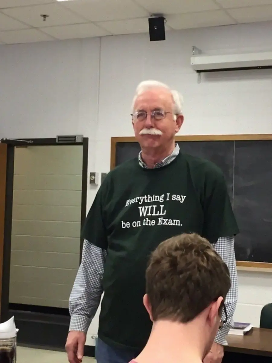 An older man with white hair and a mustache stands in a classroom wearing a dark green shirt that says, "Everything I say WILL be on the Exam." A student is seated in the foreground. A chalkboard is visible behind him.