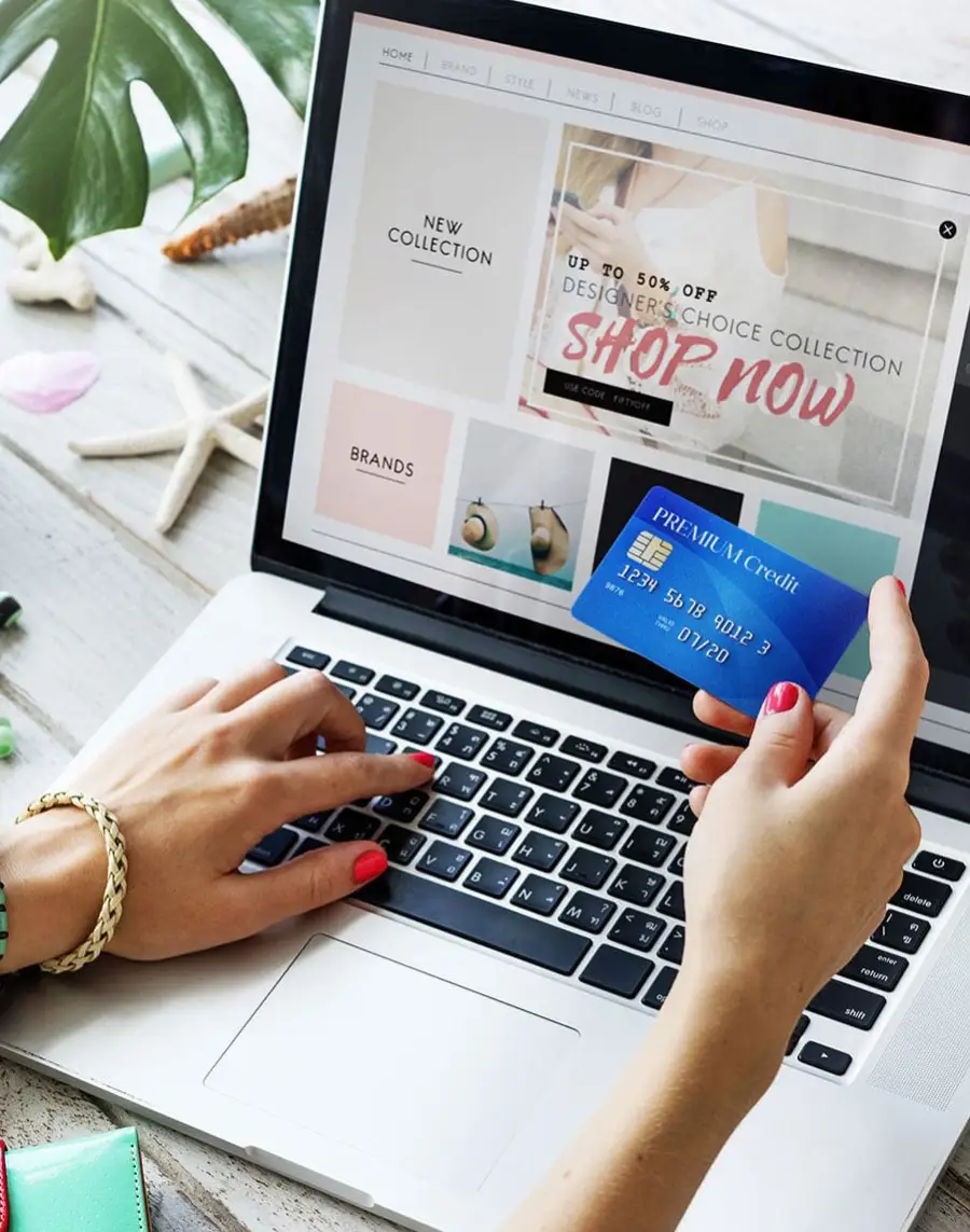 A person holding a blue credit card shops online using a laptop, with a website displaying a sale and "Shop Now" banner. A phone, notebook, and decorative items are on the white desk.