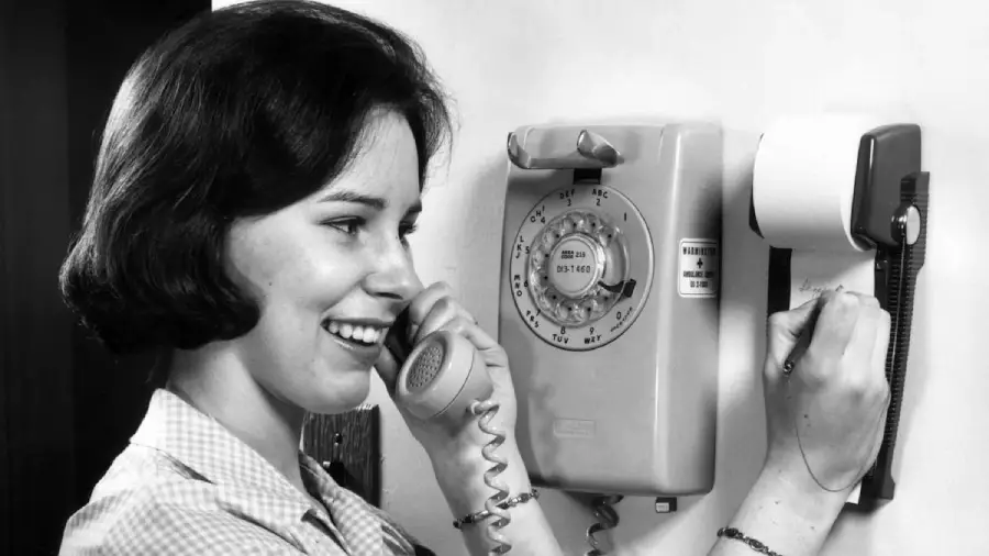 A woman smiles while talking on a rotary dial wall telephone and writes a note on a notepad attached to the wall. The image is in black and white, and the woman appears to be inside a home.
