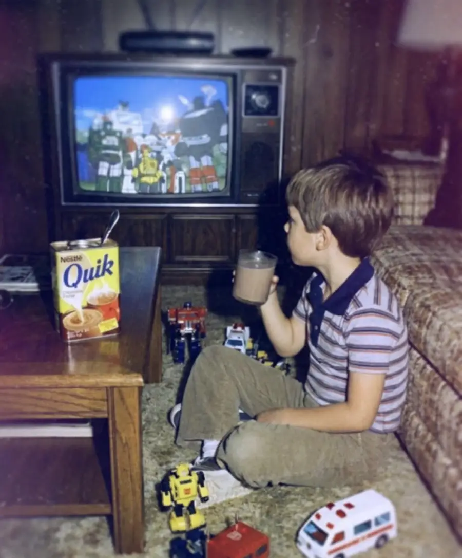 A young boy sits on the floor watching Transformers on an old TV, surrounded by toy cars and robots. He holds a glass of chocolate milk, with a Nestlé Quik box on the nearby table. The room has retro decor.