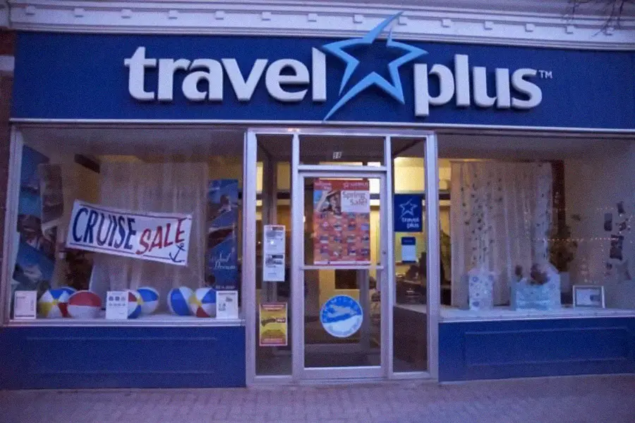 The storefront of a travel agency named "travel plus," with a large sign above the entrance and "CRUISE SALE" banner in the window, along with beach balls and travel posters displayed inside.