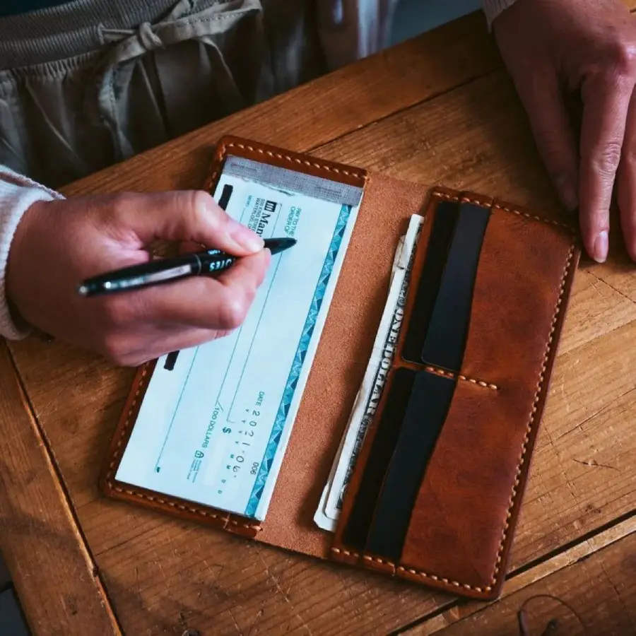A person writes on a check placed in a brown leather checkbook holder on a wooden table; cash is visible in one of the holder's pockets.
