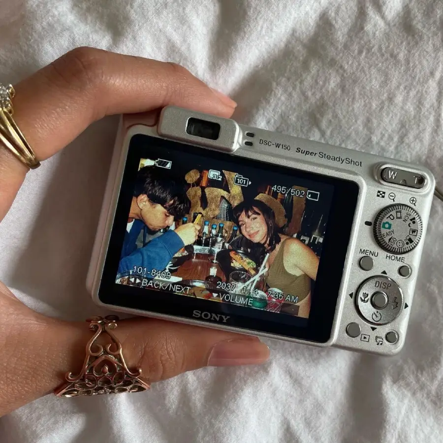 A hand holding a silver Sony digital camera, showing a photo on the screen of two people sitting at a table in a restaurant, smiling and eating food. The camera rests on a white fabric background.