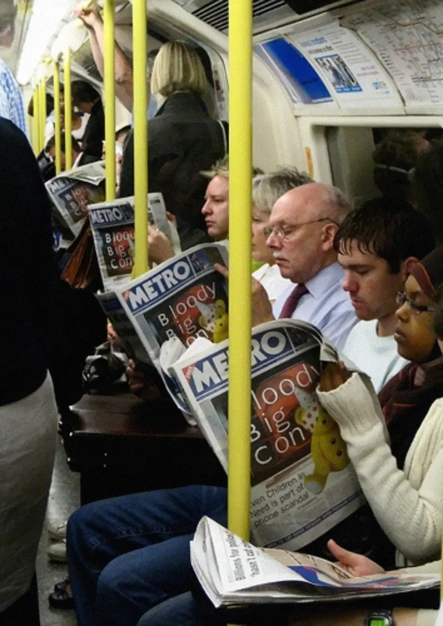 Passengers sit and stand on a crowded subway train, many reading the same Metro newspaper with the headline "Bloody Big One" and a teddy bear image on the cover. Yellow poles run through the scene.