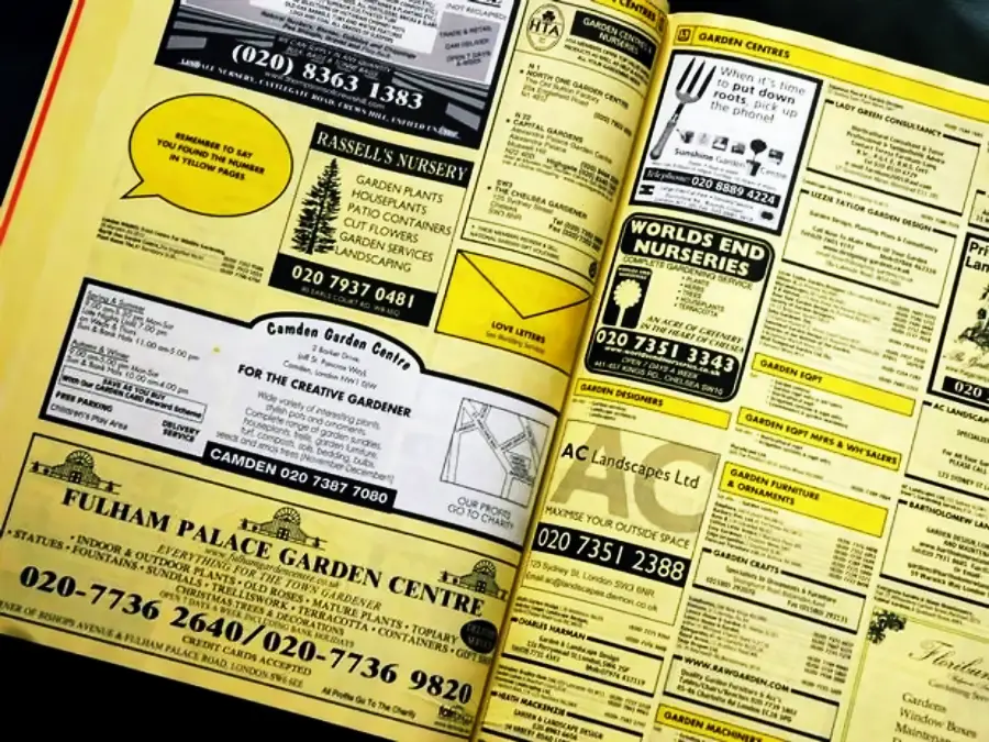 A yellow pages directory is open to a section featuring ads for various garden centers, nurseries, and landscaping services, with business names, phone numbers, and service details displayed in black and yellow text boxes.