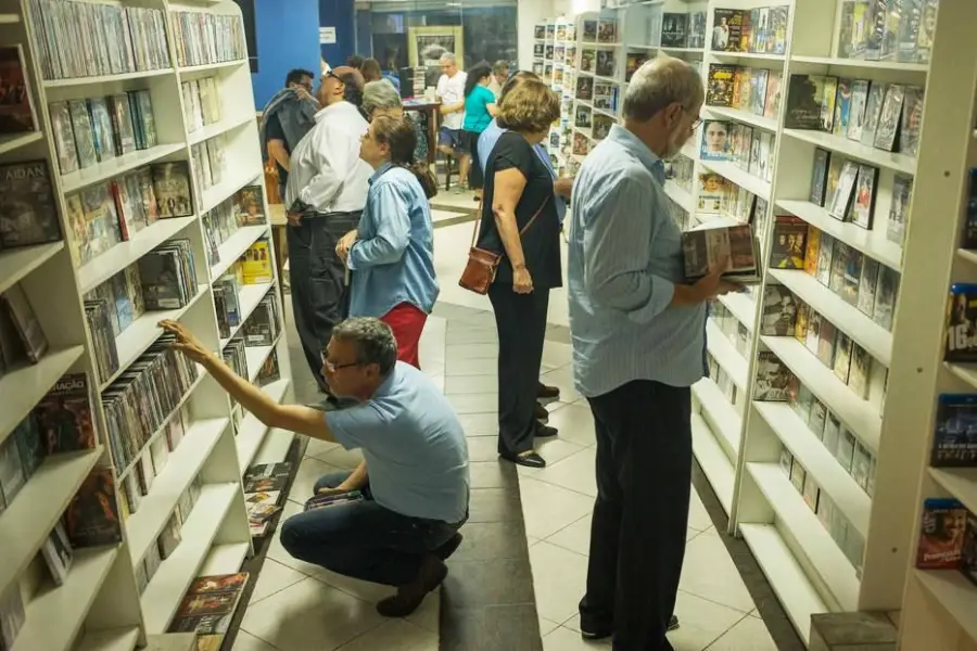 People browse DVDs on shelves in a store, with some standing and others crouching while looking at movie cases. The store is well-lit and several rows of DVDs line the white shelves.