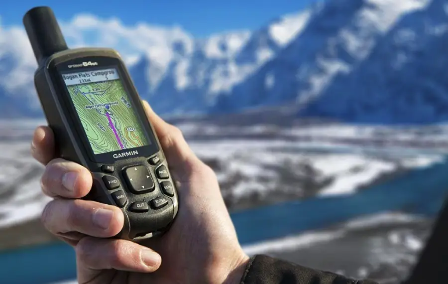 A hand holds a Garmin GPS device displaying a map with snowy mountains, a river, and a clear blue sky in the background. The scene suggests outdoor navigation or hiking in a cold, mountainous area.