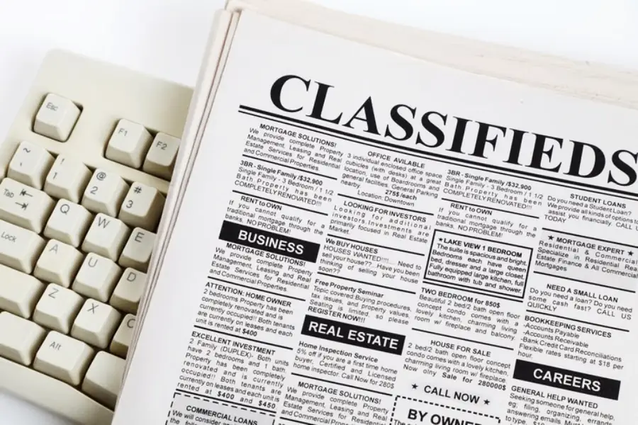 A close-up of a newspaper opened to the classifieds section, next to a beige computer keyboard. The classified ads include categories like business, real estate, and careers.