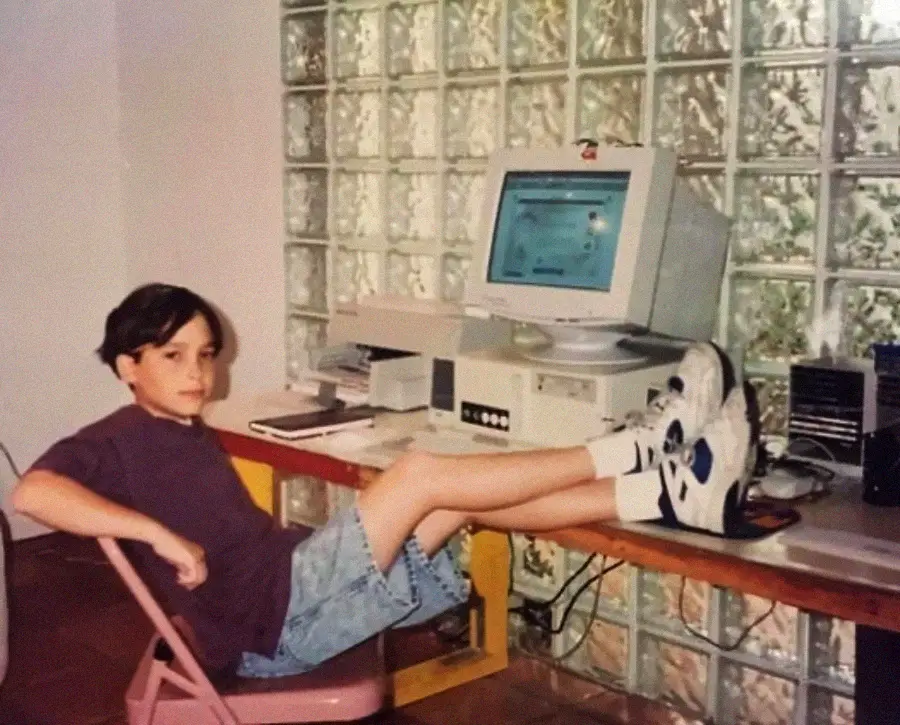 A boy in a dark T-shirt and shorts sits in a pink chair with his feet up on a desk, in front of an old desktop computer, against a wall of glass blocks.