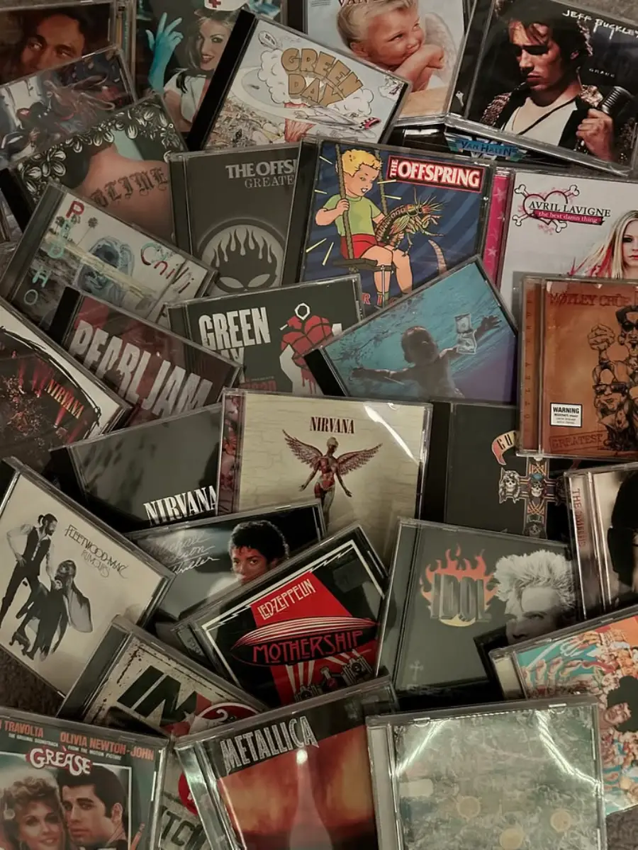 A collection of assorted CDs, featuring album covers from various artists and bands like Nirvana, Pearl Jam, Green Day, Metallica, and others, arranged overlapping each other in a scattered layout.