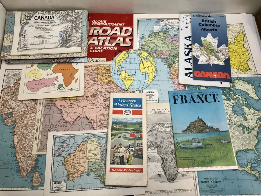 A collection of vintage road atlases, travel guides, and maps from various places including Canada, France, Alaska, British Columbia, and the Western United States, arranged on a table with a world map background.