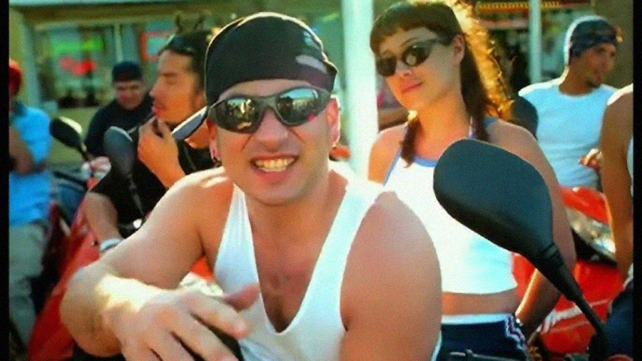 A man wearing sunglasses, a backward cap, and a white tank top smiles and gestures at the camera while a woman in sunglasses and a white tank top stands behind him. Other people and scooters are in the background.