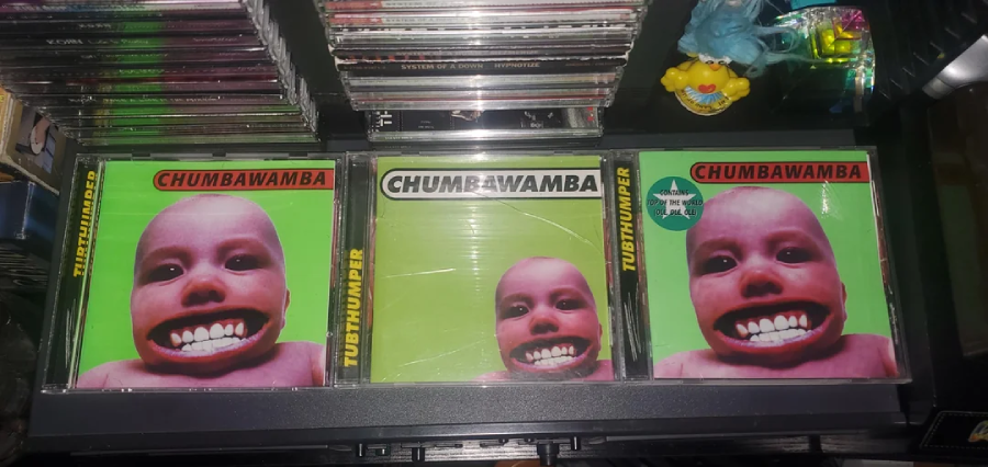 Three Chumbawamba "Tubthumper" CD cases are displayed side by side, featuring the album cover of a child's face with an exaggerated wide smile on a bright green background. CDs and figurines are nearby.