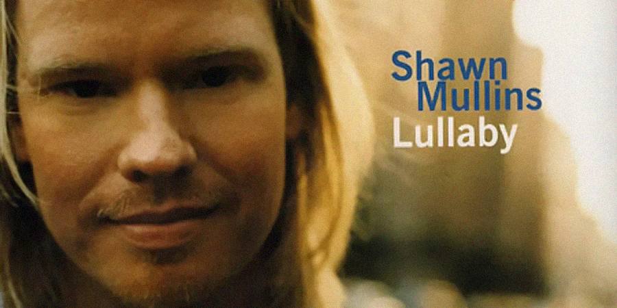 Close-up of a man with long blond hair and a beard, smiling softly. The text next to him reads "Shawn Mullins Lullaby" in blue and white letters. The background is blurred.