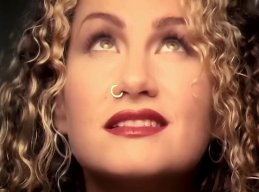 A woman with curly blonde hair and a gold nose ring looks upward, smiling softly. She has light eyes, red lipstick, and is shown in close-up with a warm, soft-focus background.
