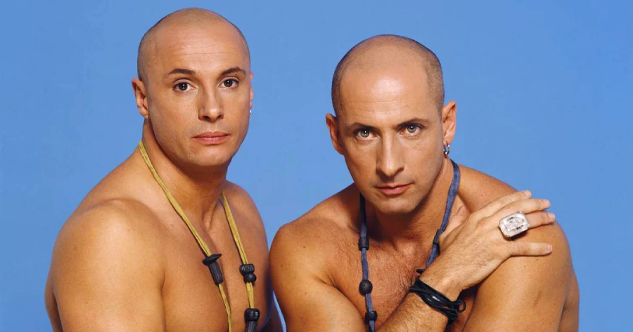 Two bald men with muscular builds pose shirtless against a blue background. Both wear necklaces made of chunky black beads, and one man rests his hand on the other's shoulder, displaying a large ring.