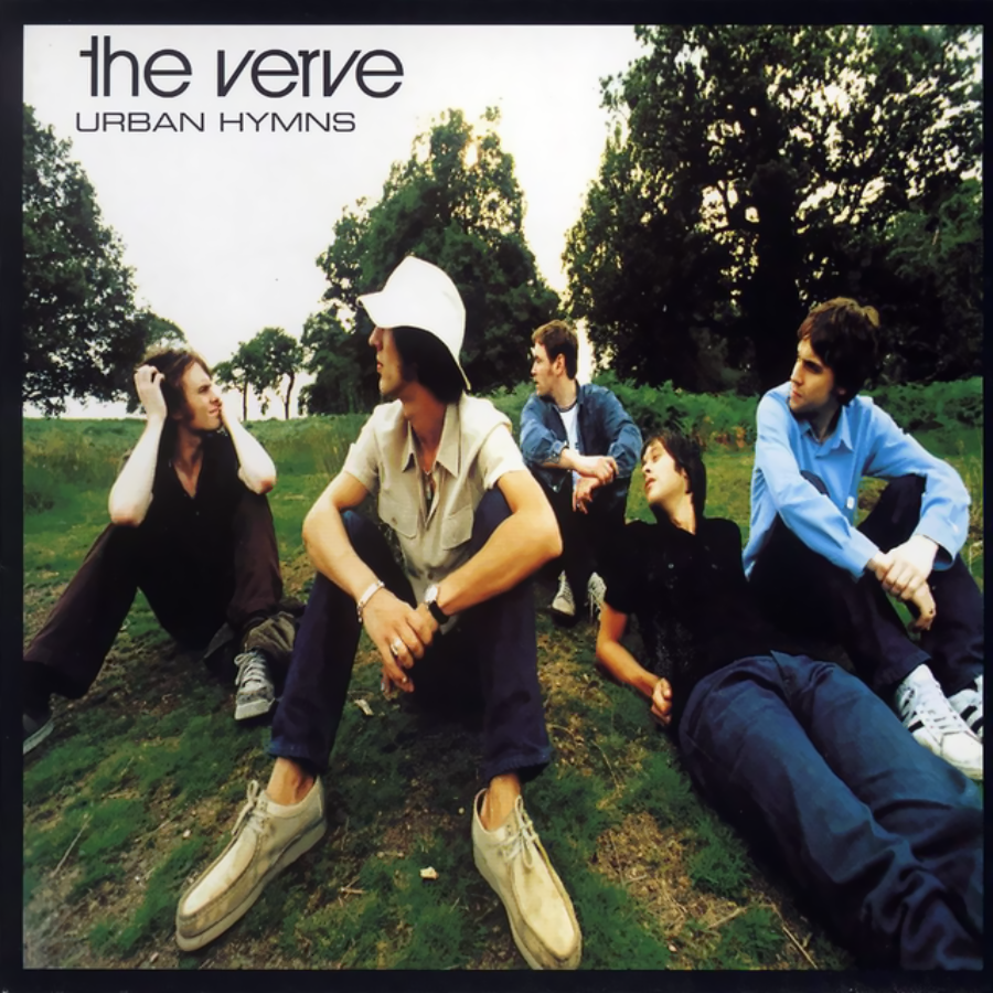 Five young men sit on grass in a park, surrounded by trees, casually talking. "The Verve" and "Urban Hymns" are written in the top left corner, indicating an album cover.