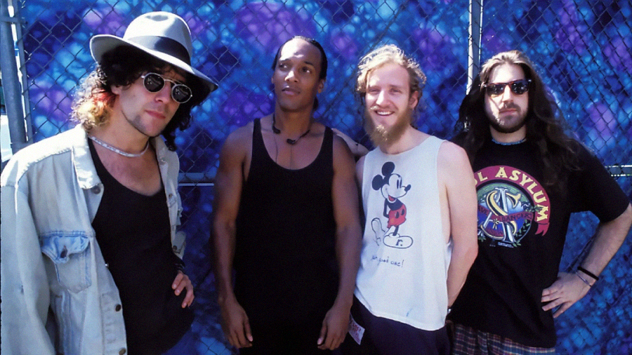 Four men stand in front of a blue graffiti wall and chain-link fence. They wear casual 1990s clothes, including tank tops, a hat, and sunglasses, and all have relaxed, friendly expressions.