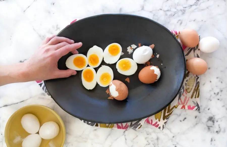 A hand reaches for a black plate holding several halved boiled eggs and some eggs with cracked shells, resting on a patterned cloth. Whole eggs and a yellow bowl with eggs are nearby on a marble surface.