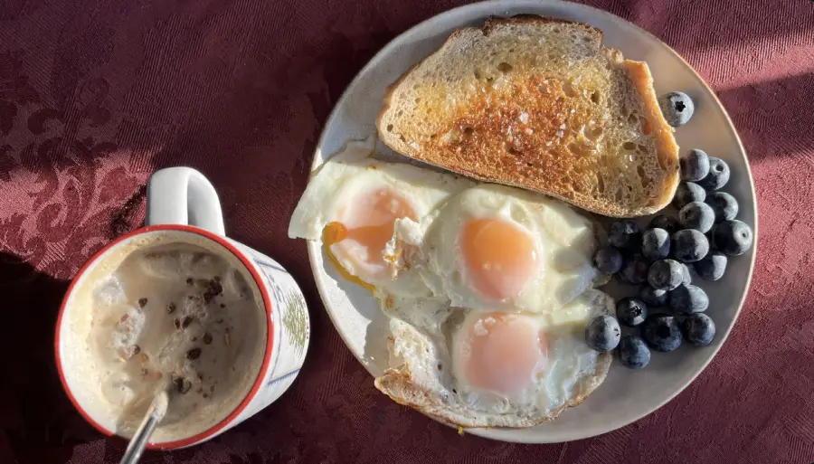 A breakfast plate with three sunny-side-up eggs, two slices of buttered toast, and a handful of blueberries, next to a mug of frothy coffee on a red tablecloth.