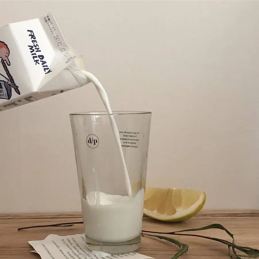 A carton labeled "Fresh Daily Milk" pours milk into a clear glass on a wooden surface. Nearby are a lemon wedge, a green plant stem, and a piece of paper with text. The background is plain and light-colored.
