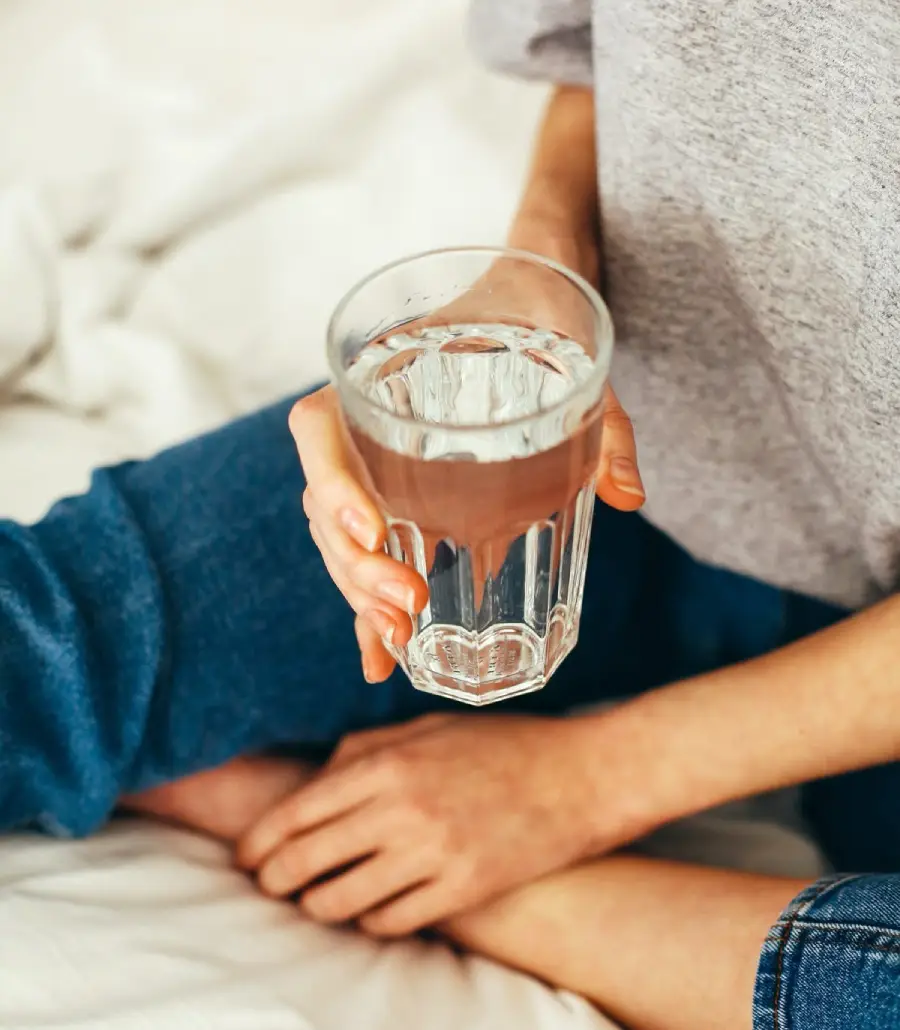 A person sitting cross-legged on a bed holds a clear glass of water in one hand, wearing blue jeans and a light gray shirt. The background is softly blurred.