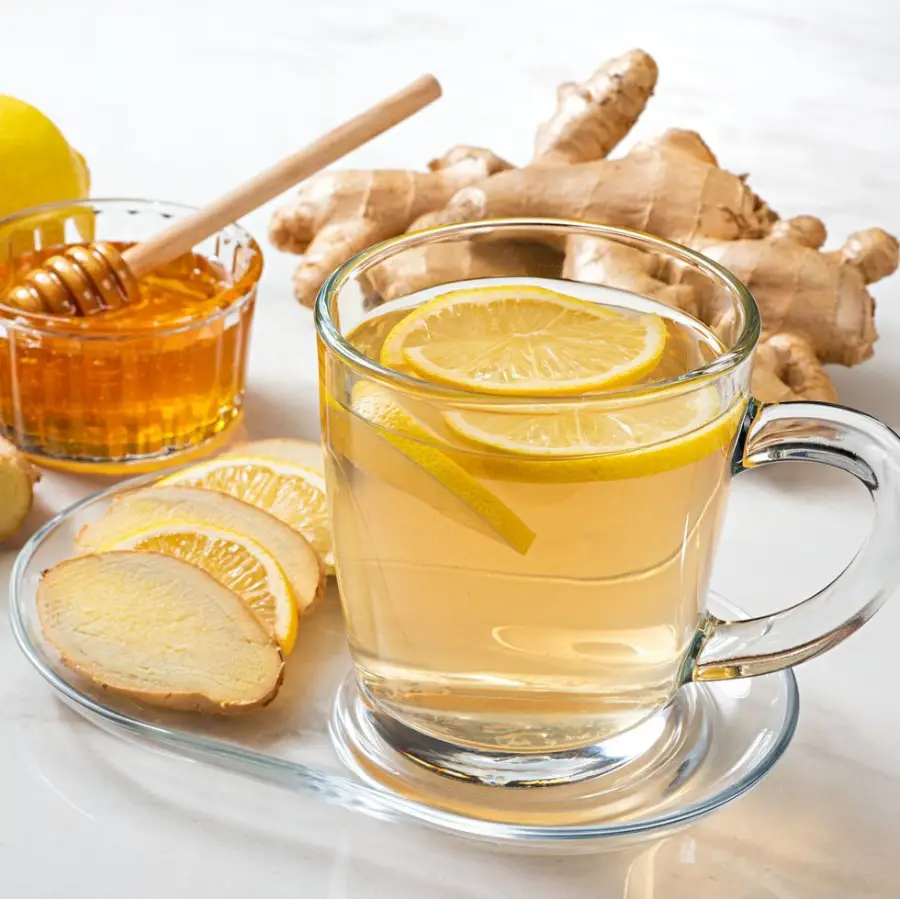 A glass cup of hot lemon ginger tea with lemon slices, fresh ginger, a dish with lemon and ginger slices, and a small bowl of honey with a honey dipper on a white surface.