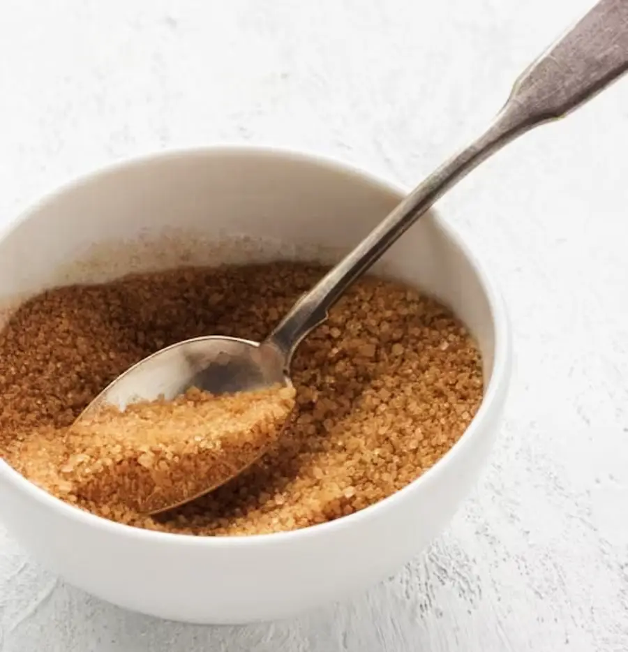 A white bowl filled with coarse brown sugar, with a metal spoon scooping some of the sugar, placed on a light textured surface.