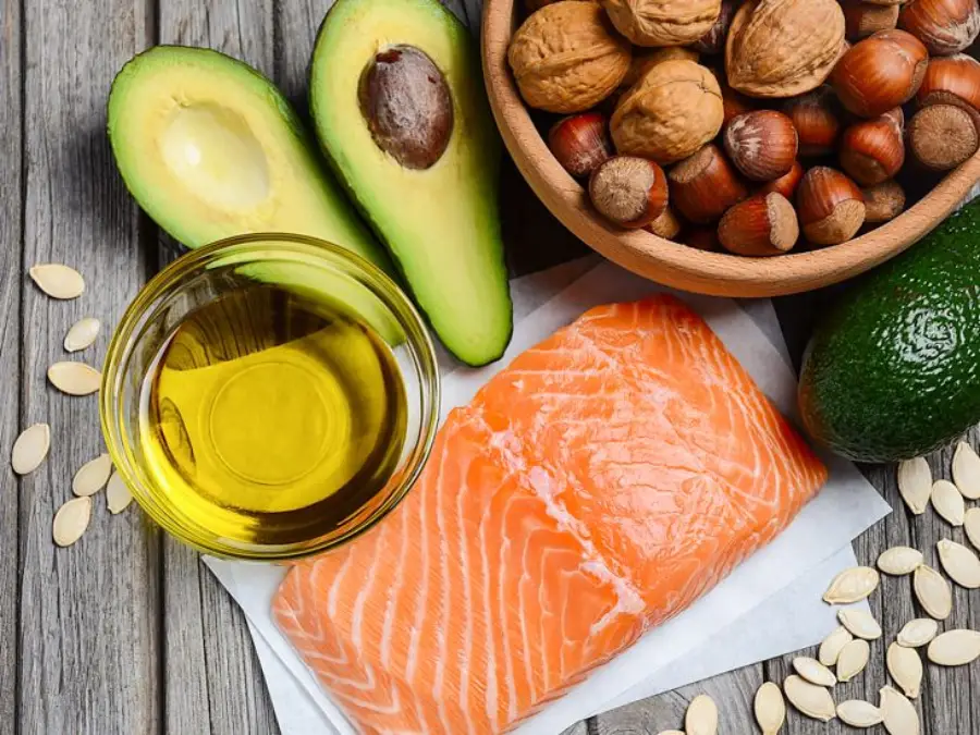 A wooden table displays a raw salmon fillet, a bowl of mixed nuts, a halved avocado, a whole avocado, a small glass bowl of oil, and scattered pumpkin seeds.