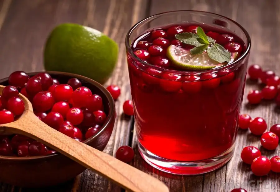 A glass of cranberry juice garnished with a lime slice and mint, sits next to a bowl of fresh cranberries with a wooden spoon, on a rustic wooden table. Half a lime is also visible in the background.