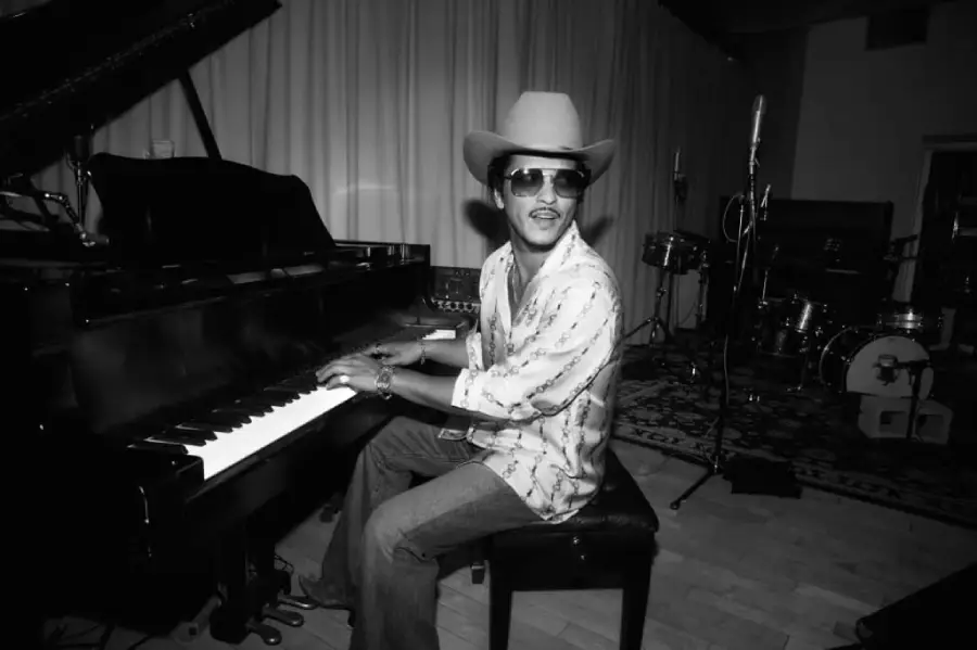 A man wearing a cowboy hat, sunglasses, and a patterned shirt plays a grand piano in a music studio with drums and microphones in the background. The photo is in black and white.