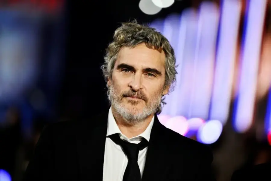 A man with wavy gray hair and a beard, wearing a black suit, white shirt, and loose black tie, stands against a blurred background with colorful lights.