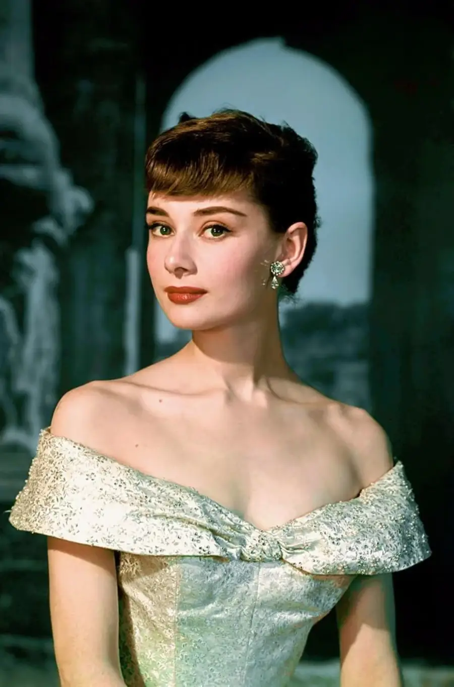 A woman with short dark hair, wearing an off-the-shoulder, silver gown and earrings, poses gracefully against a dark, blurred background with architectural elements.