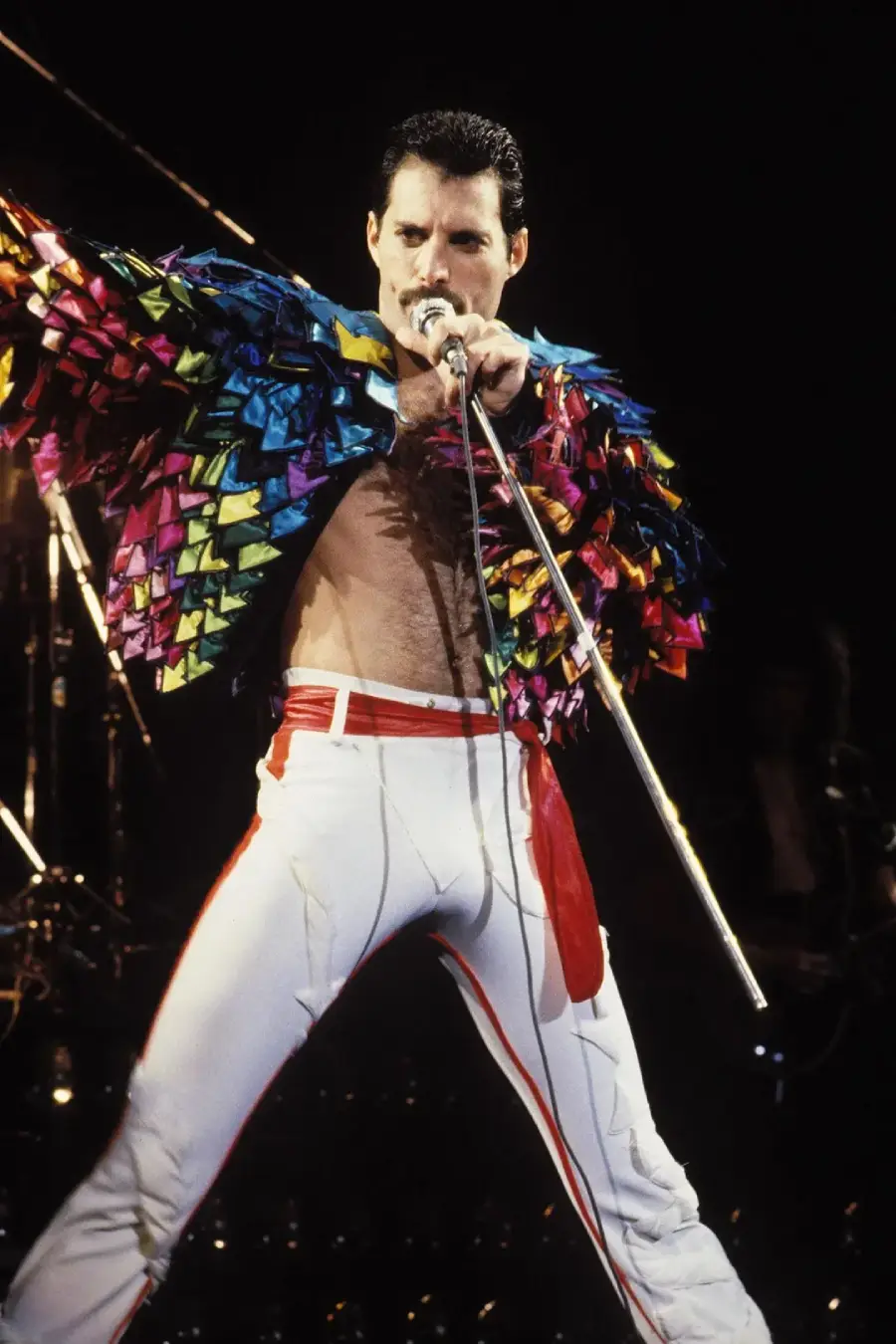A performer in a colorful, feathered jacket and white pants sings passionately into a microphone on stage, striking a dramatic pose under stage lights.