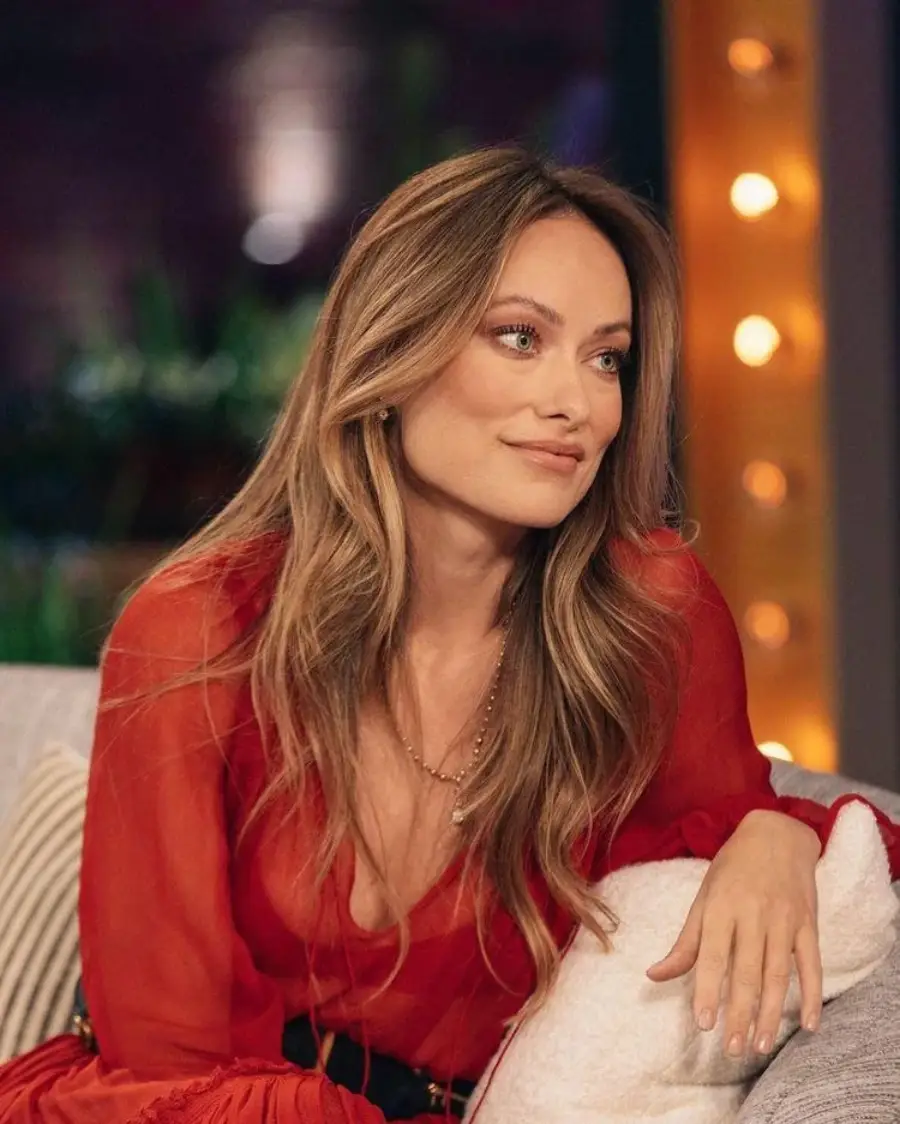 A woman with long, wavy light brown hair wearing a red blouse sits on a couch, smiling softly and looking to the side. Warm lights and a blurred background create a cozy atmosphere.