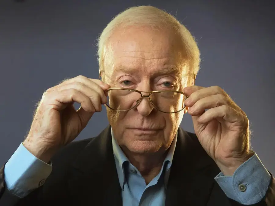 An elderly man with white hair and a blue shirt adjusts his glasses, looking directly at the camera with a serious expression. The background is plain and softly lit.