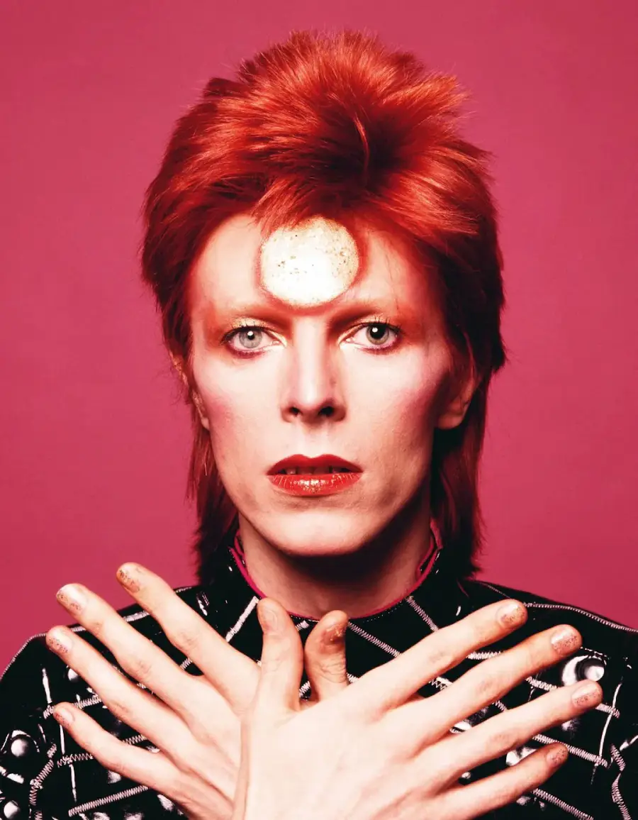 A person with bright red hair and dramatic makeup poses against a pink background, hands crossed over their chest with painted nails, wearing a patterned black outfit and a gold circle on their forehead.