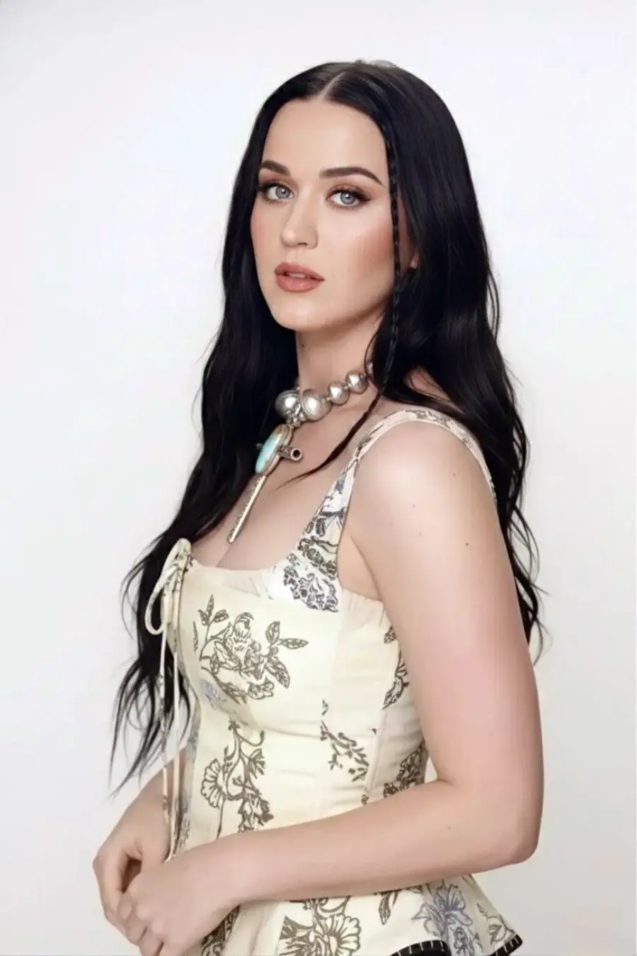 A woman with long dark hair and blue eyes poses against a plain background. She is wearing a cream-colored, floral-printed top and a statement necklace with large beads and turquoise accents. She gazes confidently at the camera.