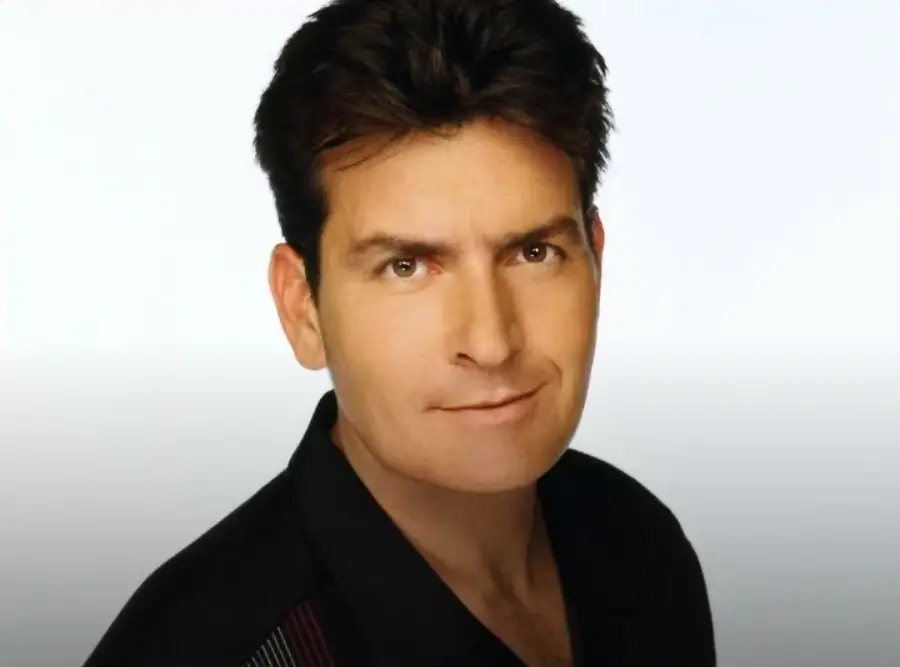 A man with short dark hair and fair skin, wearing a black collared shirt, looks toward the camera with a slight smile against a plain light background.
