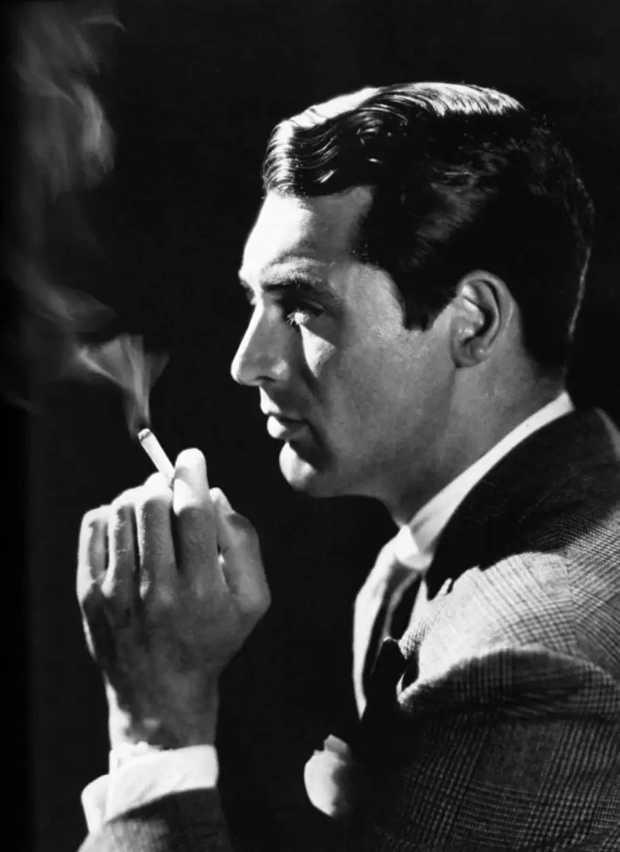 Black and white photo of a man in a suit, shown in profile, holding a lit cigarette near his face with smoke rising, set against a dark background.