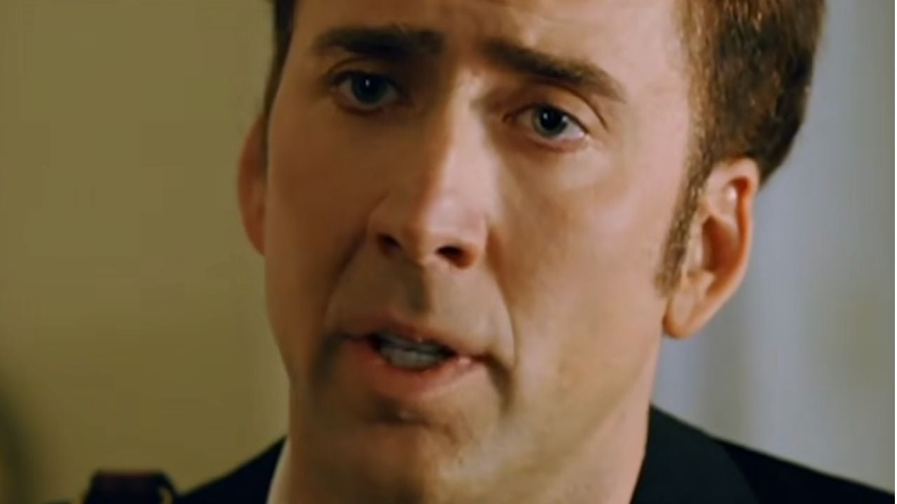 A close-up of a man with short brown hair, light skin, and blue eyes, looking slightly to the side with a serious expression. He is dressed formally and appears to be speaking or listening intently.