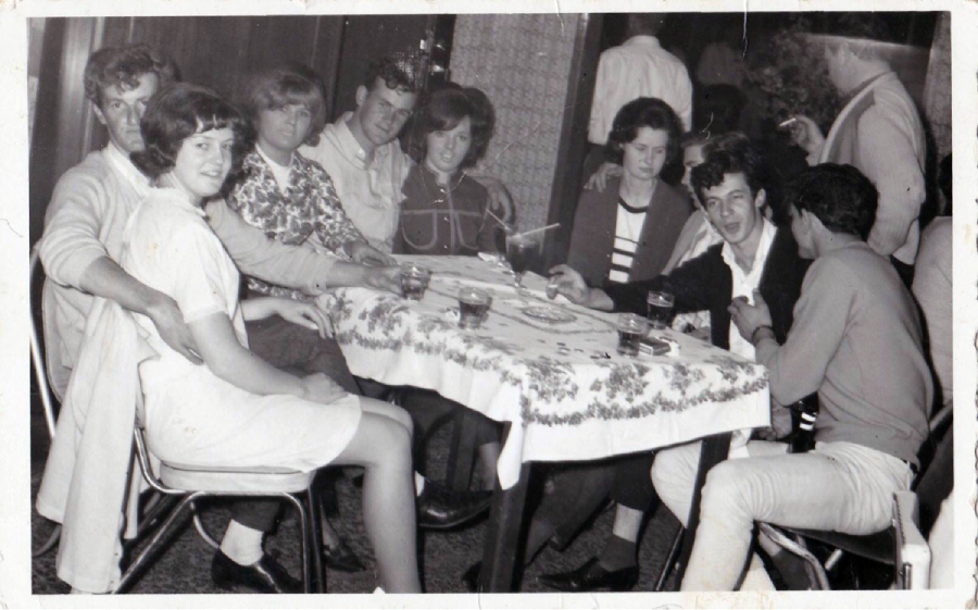 A black-and-white photo of eight young people sitting around a table with drinks, chatting and smiling. The table is covered with a floral tablecloth, and additional people are visible in the background.