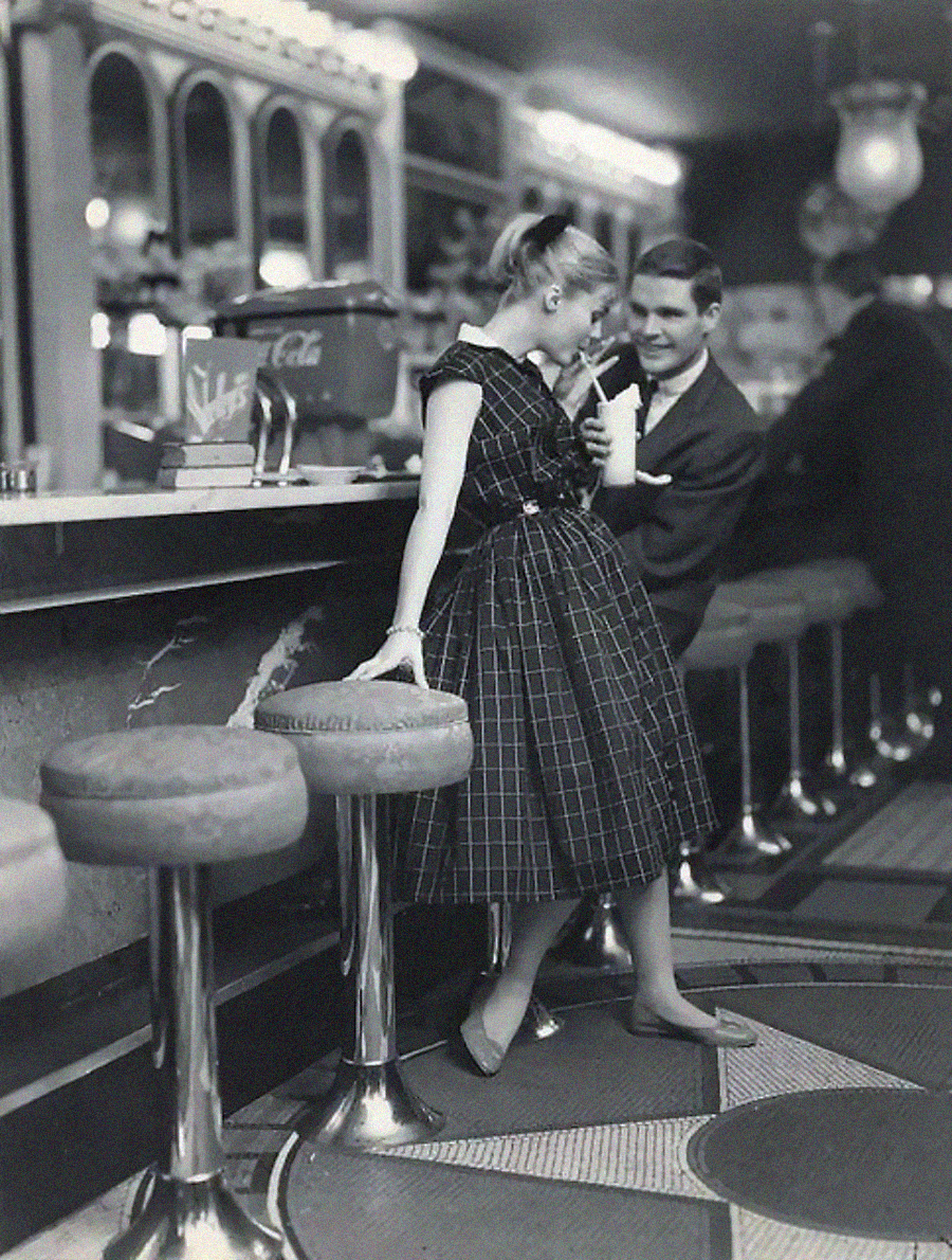 A young woman in a plaid dress sips a drink at a soda fountain counter while a young man in a suit leans beside her. The retro diner has round stools, a checkerboard floor, and vintage decor.