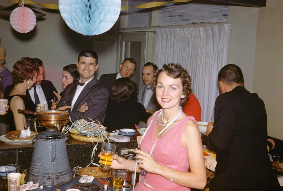 A woman in a pink dress smiles at the camera while holding a drink at a lively indoor party, with several people socializing and decorations hanging above. Food and drinks are spread on the counter.