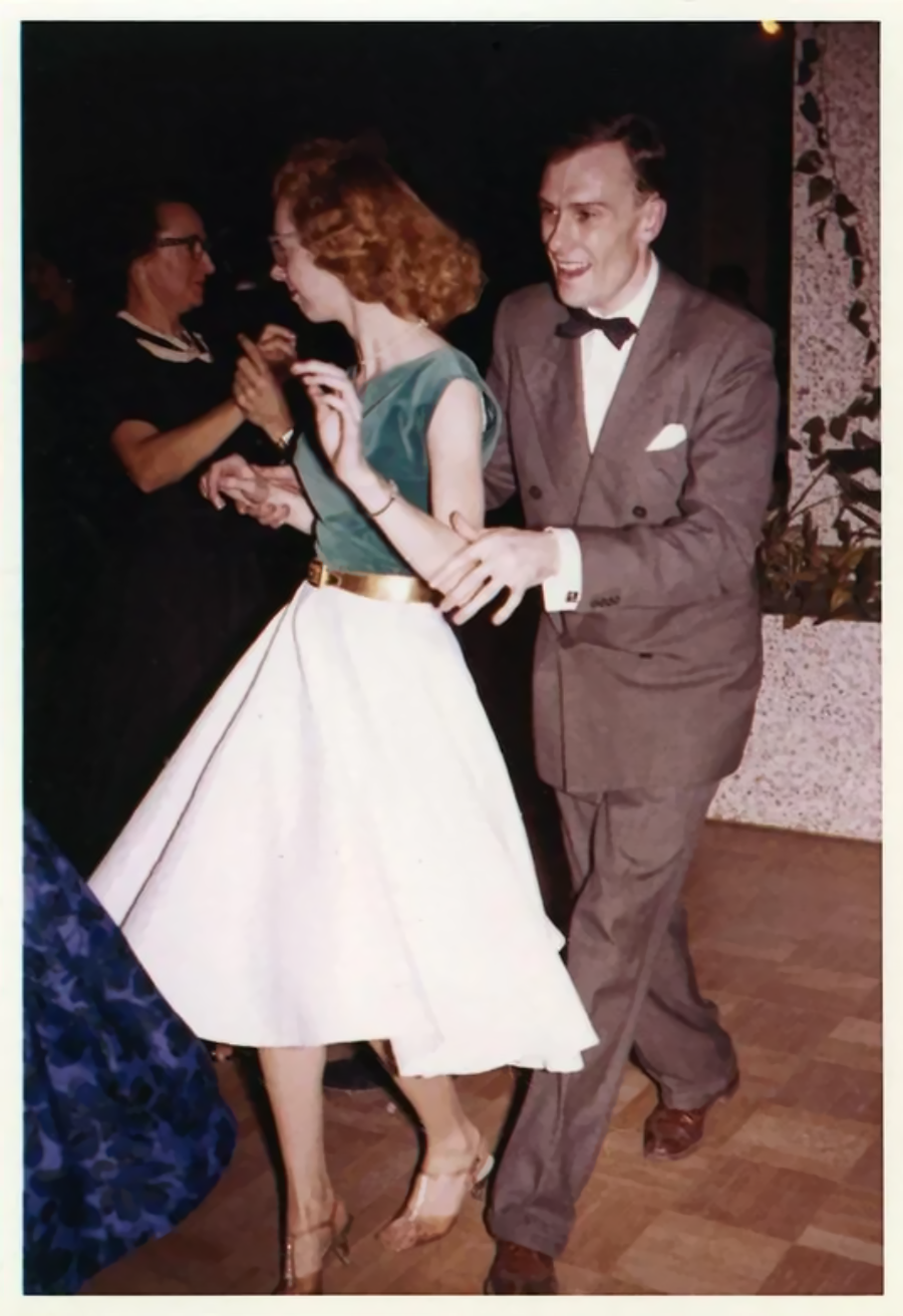 A man in a gray suit and bow tie dances with a woman in a green top and white skirt on a wooden floor, surrounded by other people at a formal event.